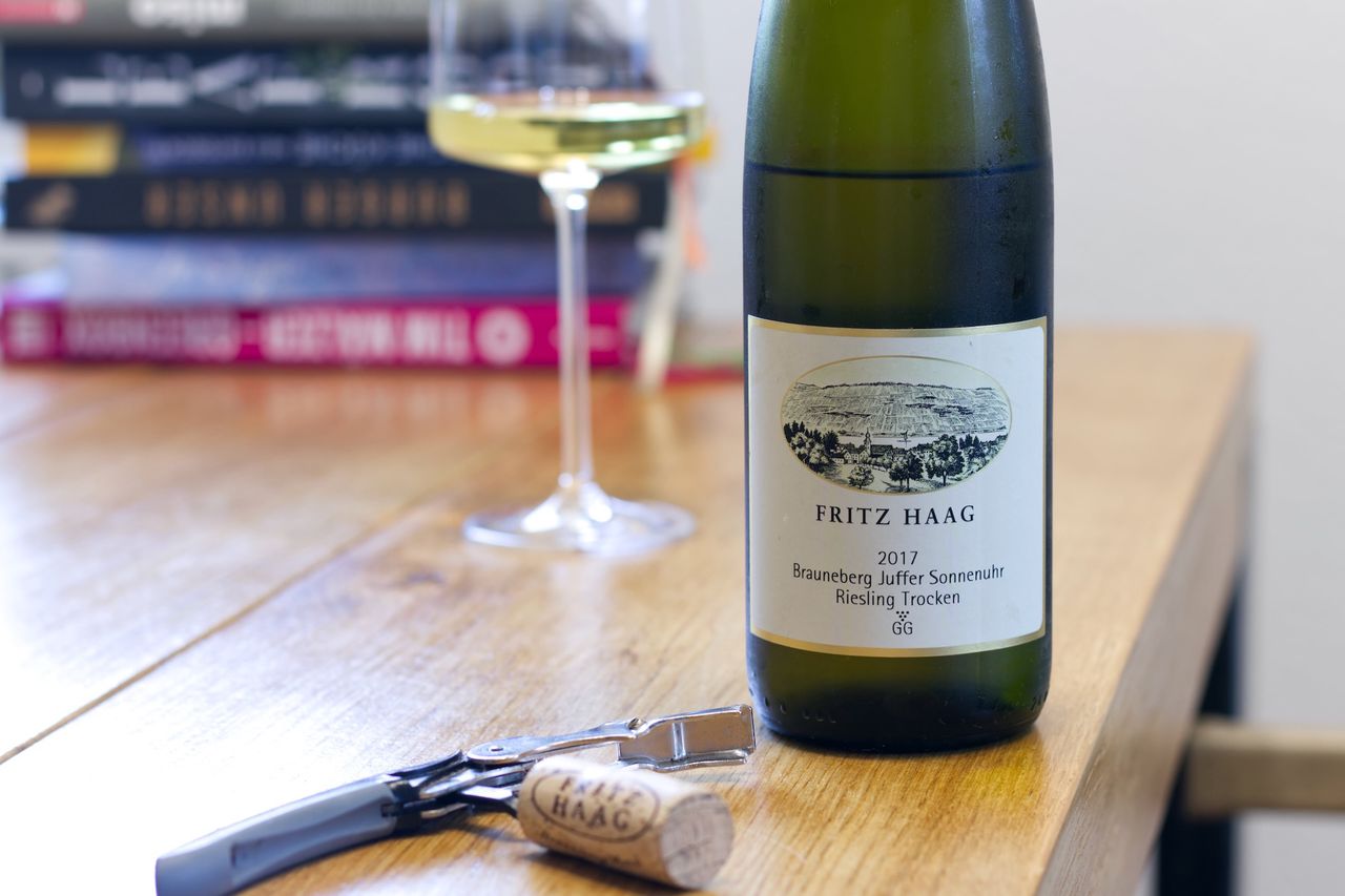 Eine Flasche Brauneberg Juffer Sonnenuhr Großes Gewächs 2017 vom Weingut Fritz Haag auf einem Holztisch, mit einem Weinglas und Büchern im Hintergrund sowie einem Korken und einem Korkenzieher im Vordergrund.