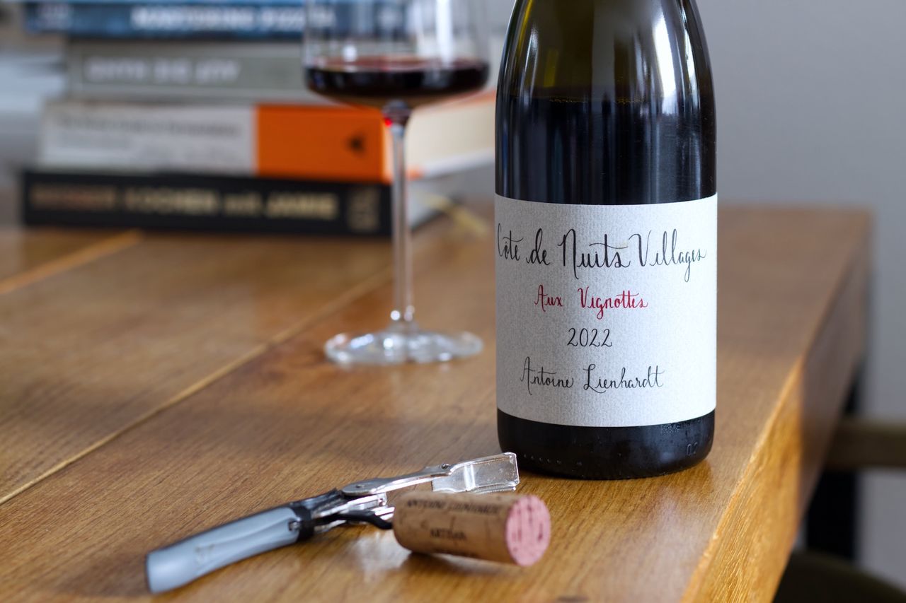 Eine Flasche Wein von Antoine Lienhardt aus dem Jahr 2022. Auf dem Etikett steht in geschwungener Schrift Côte de Nuits Villages und Aux Vignottes. Im Hintergrund sind ein Weinglas und Bücherstapel zu sehen. Vor der Flasche liegt der Korken der Flasche am Kellnermesser.