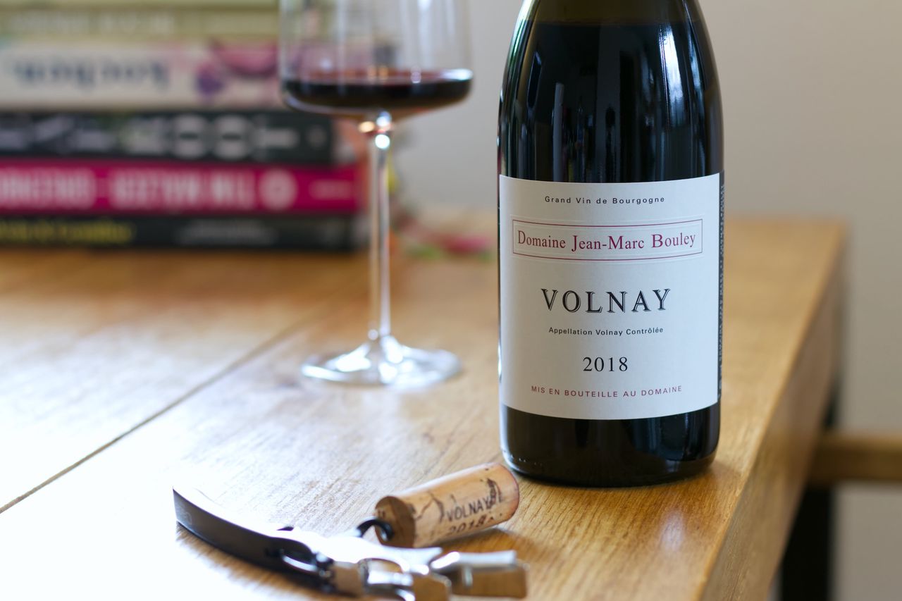 Eine Flasche Volnay 2018 von der Domaine Jean-Marc Bouley auf einem Holztisch. Im Hintergrund steht ein gefülltes Weinglas vor einem Stapel Kochbücher, im Vordergrund liegt ein Korken an einem Kellnermesser.