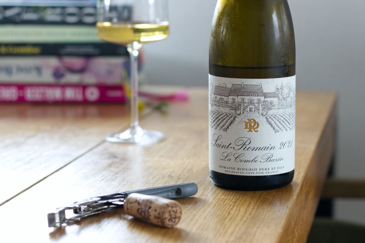 Eine Flasche Saint-Romain La Combe Bazin 2021 von der Domaine Denis Rougeot auf einem Holztisch. Im Hintergrund steht ein gefülltes Weinglas vor einem Stapel Kochbücher, im Vordergrund liegt der passende Korken an einem Kellnermesser.