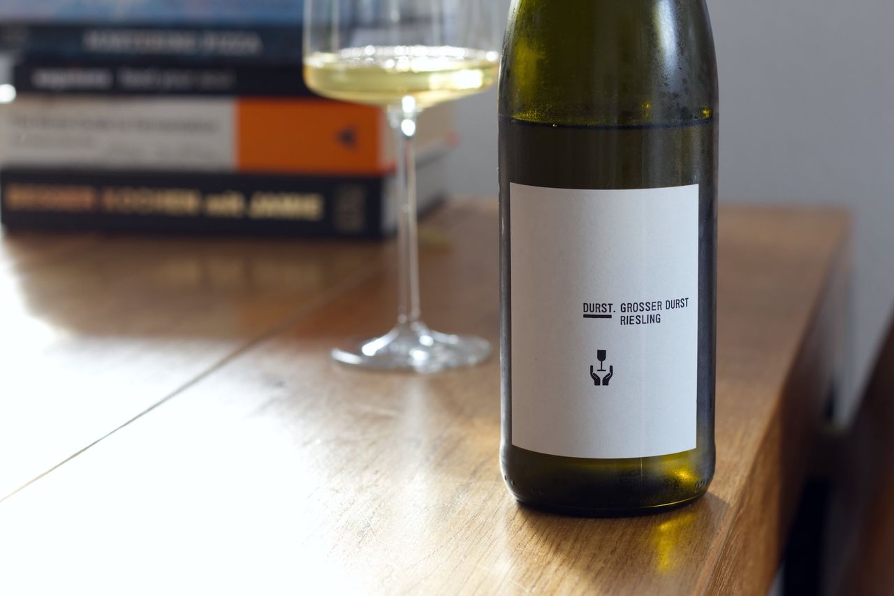 Eine Flasche Riesling Grosser Durst 2018 von Andreas Durst auf einem Holztisch. Im Hintergrund sind ein Weinglas und Bücherstapel zu sehen.