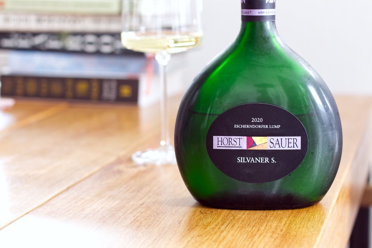 Eine Bocksbeutel-Flasche Wein Silvaner S aus der Lage Escherndorfer Lump von Horst Sauer auf einem Holztisch. Im Hintergrund sind ein Weinglas und Bücherstapel zu sehen.