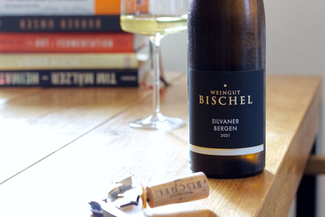 Eine Flasche Silvaner Bergen 2021 vom Weingut Bischel auf einem Holztisch. Im Hintergrund steht ein gefülltes Weinglas vor einem Stapel Kochbücher, im Vordergrund liegt der passende Korken an einem Kellnermesser.