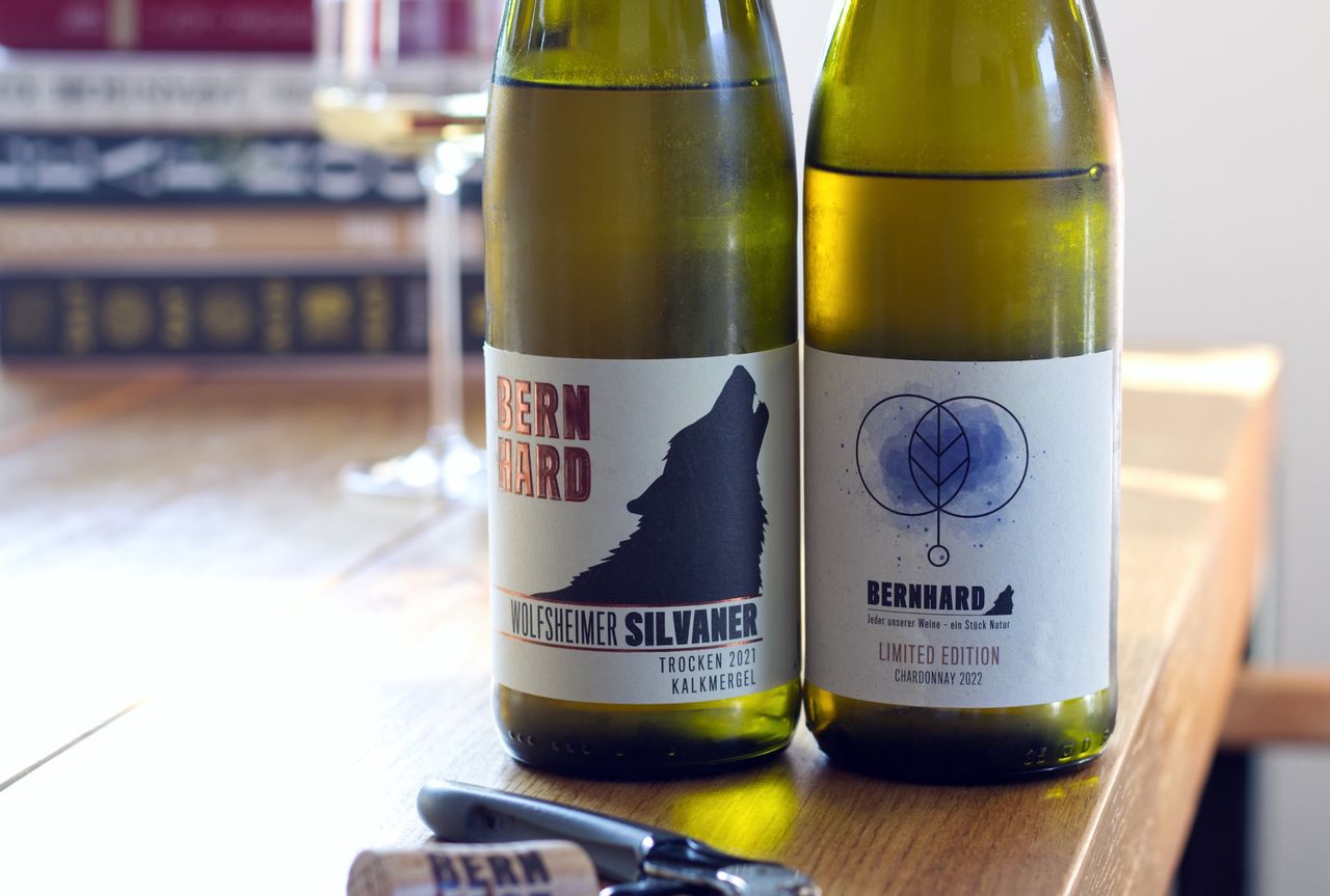 Auf einem Holztisch stehen zwei Flaschen Wein vom Weingut Bernhard. Ein Chardonnay in einer limitierten Edition und ein Silvaner. Im Hintergrund sind ein Weinglas und ein Bücherstapel zu sehen. Vor den Flaschen liegt Korken und Kellnermesser.