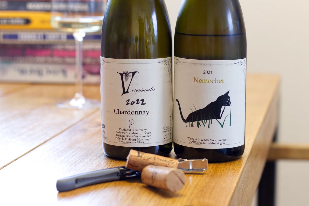 Auf einem Holztisch stehen zwei Flaschen Wein vom Weingut Vorgrimmler. Ein Chardonnay 2022 und ein Nemochet 2021 mit einer Katze auf dem Etikett. Im Hintergrund sind ein Weinglas und ein Bücherstapel zu sehen.