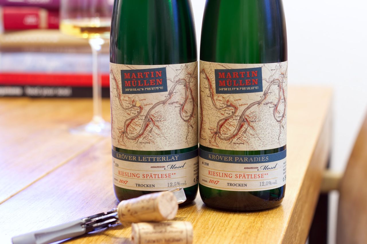 Auf einem Holztisch stehen zwei Flaschen Wein vom Weingut Martin Müllen. Eine Riesling Spätlese aus dem Kröver Paradies, eine Spätlese aus dem Kröver Letterlay. Die Etiketten zeigen eine Karte der Mosel. Im Hintergrund sind ein Weinglas und ein Bücherstapel zu sehen.